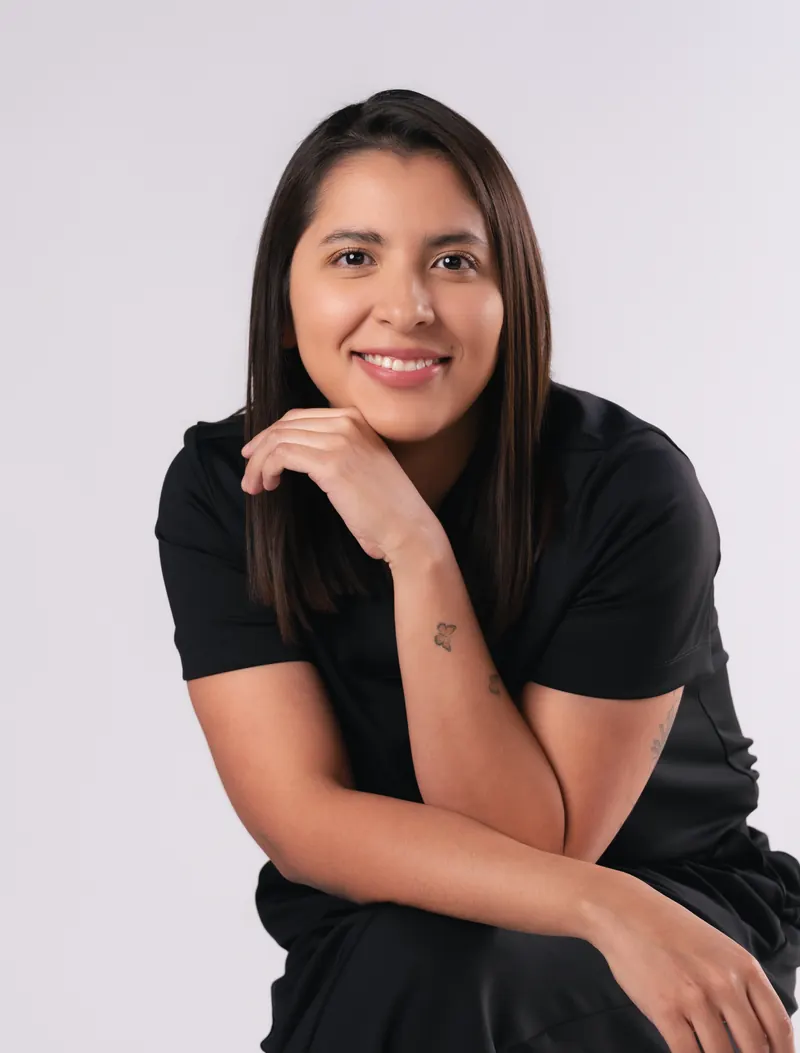 La Dra. Lourdes Alanis Naranjo, sentada con uniforme quirúrgico negro, apoya la barbilla en la mano y sonríe.