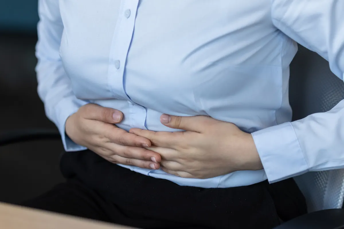 Mujer con blusa clara se sujeta el abdomen con ambas manos por el dolor.