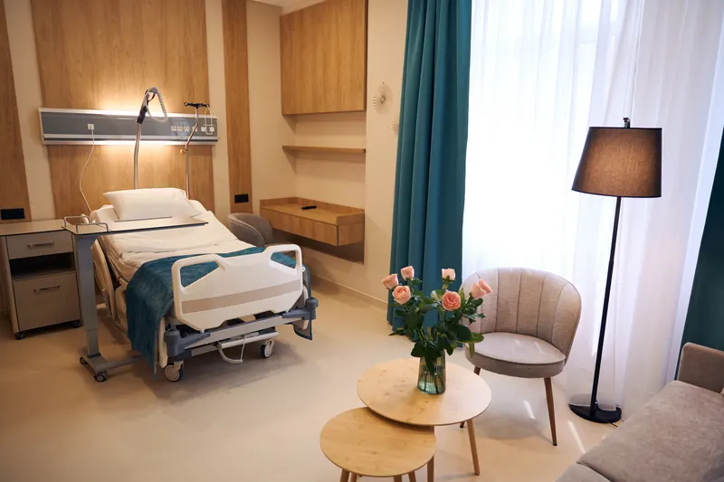 Habitación de hospital con cama, sillón, lámpara y un florero con rosas, iluminada con luz natural.